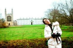 A young Chinese woman warmly dressed and holding a camera poses and pulls a funny face with a cathedral and stately home in the background.